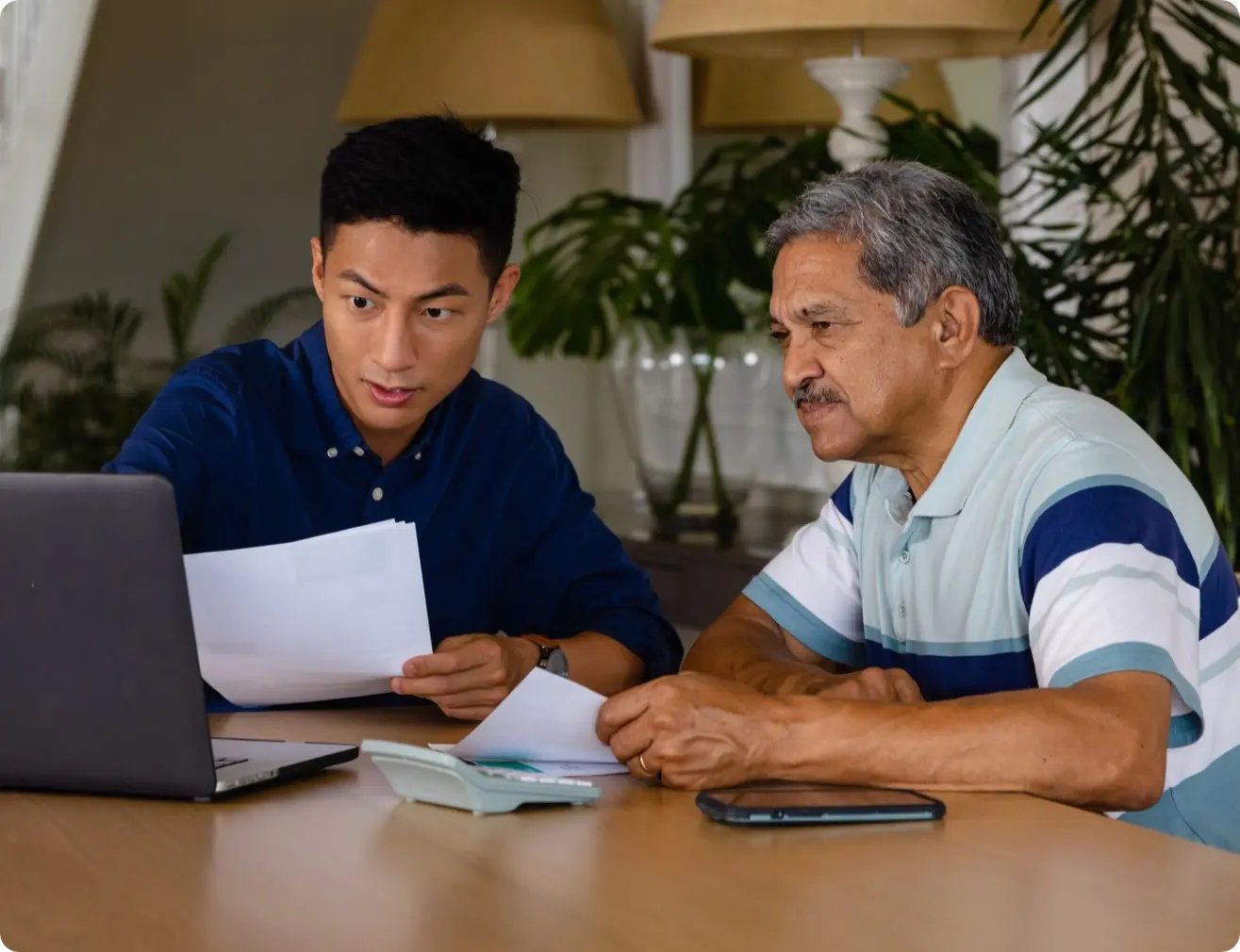 A young man and his parent sit together at a table, looking intently at a laptop screen with focused, concerned expressions while holding paper documents and working through college applications.