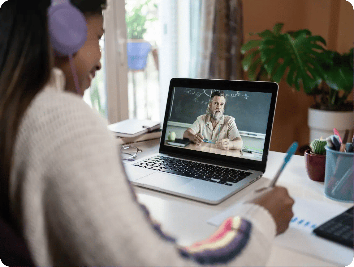Student wearing headphones taking notes during a video call with an online SAT tutor on a laptop. Student wearing headphones taking notes during a video call with an online SAT tutor on a laptop.